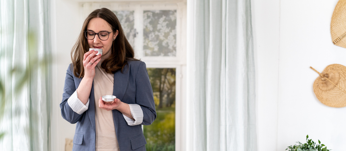 Frau in blauem Blazer und hellem Oberteil hält ein kleines Gefäß an die Nase in einem hellen Raum mit Pflanzen und Wanddekoration