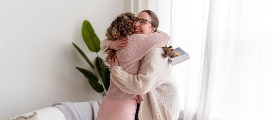 Zwei Frauen umarmen sich herzlich in einem hellen Raum, eine hält eine Geschenkbox hinter dem Rücken