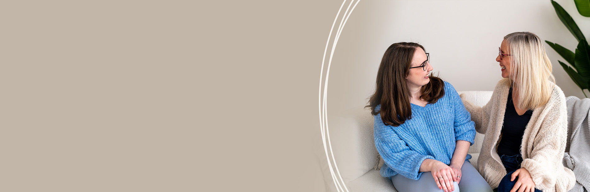 Zwei Frauen sitzen nebeneinander, eine trägt einen blauen Pullover, die andere eine helle Strickjacke, beide blicken sich an.