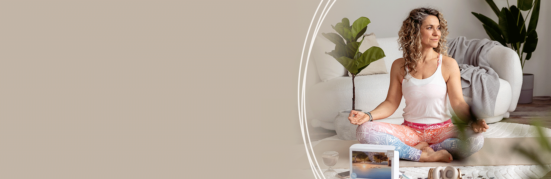 Frau sitzt im Schneidersitz auf einem Teppich, trägt weiße Tanktop und bunte Hose, umgeben von Pflanzen und Kissen.