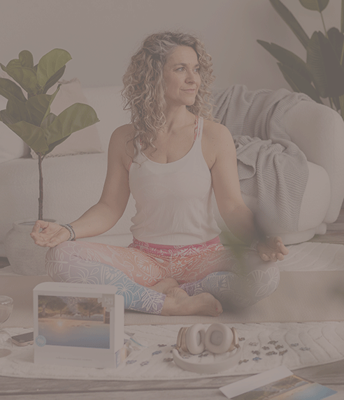 Frau sitzt im Schneidersitz auf einem Teppich, trägt weiße Tanktop und bunte Hose, umgeben von Pflanzen und Kissen.