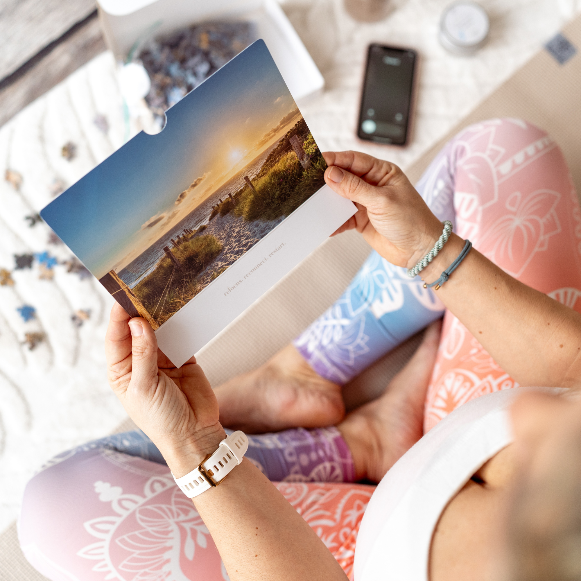 Frau sitzt im Schneidersitz auf buntem Yogamattenmuster und hält eine Postkarte mit Sonnenuntergang über Dünen