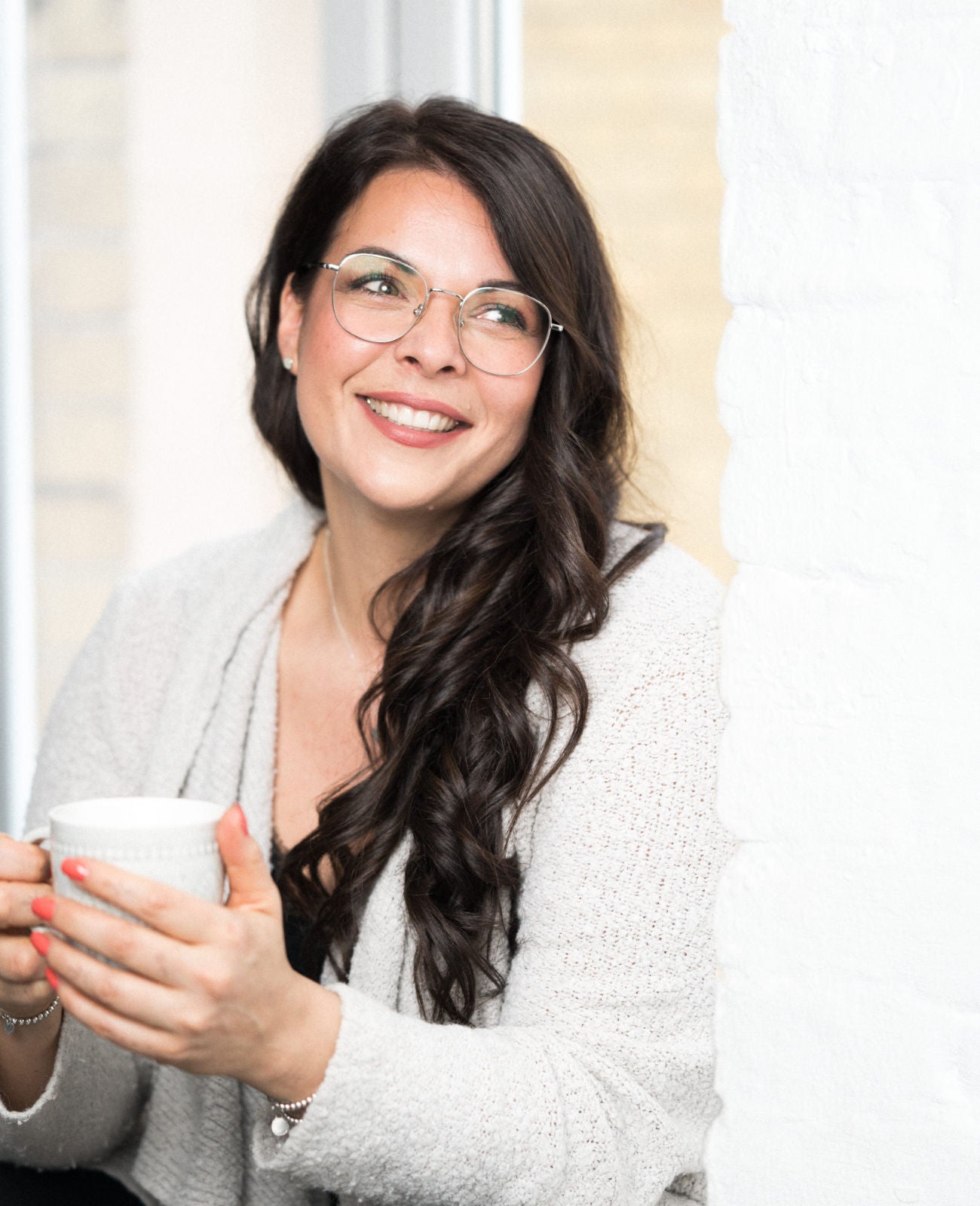 Frau mit langen, dunklen, gewellten Haaren lehnt an weißer Wand und hält eine weiße Tasse in beiden Händen.