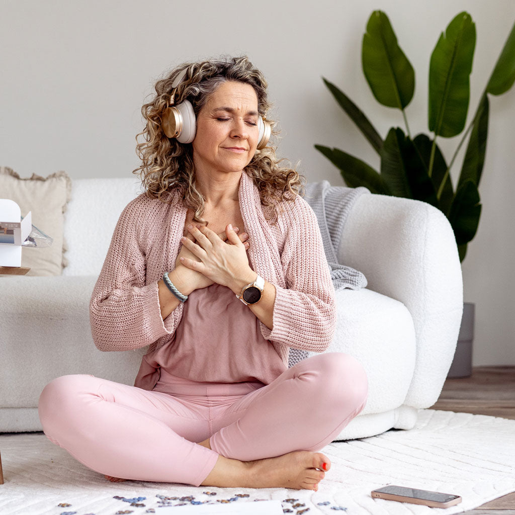 Frau sitzt im Schneidersitz auf einem Teppich vor einem weißen Sofa, trägt rosa Kleidung und goldene Kopfhörer, Hände auf der Brust.