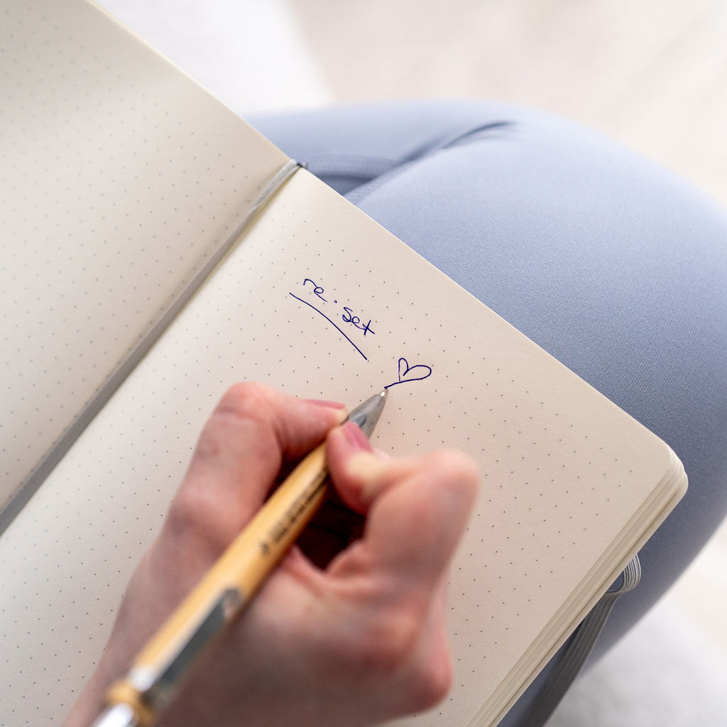 Hand schreibt mit silbernem Kugelschreiber auf ein gepunktetes Notizbuch, Text 'reset' und ein Herzsymbol sichtbar.