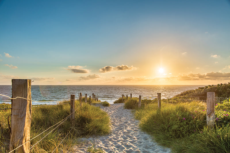 Sandiger Weg zwischen Dünen mit Gras und Holzzäunen führt zum Meer bei Sonnenuntergang
