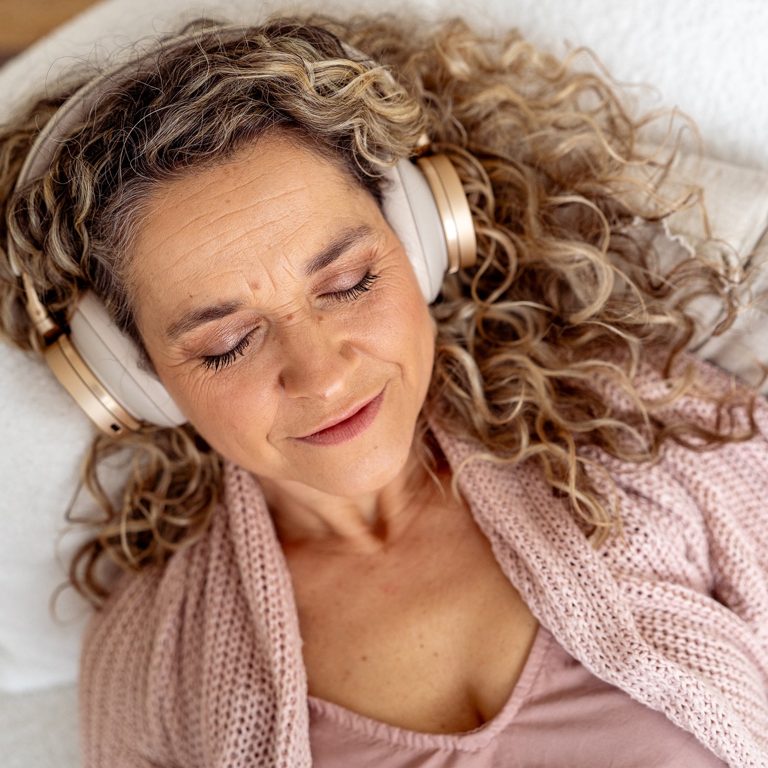 Frau mit lockigem Haar und Kopfhörern entspannt mit geschlossenen Augen auf einem Kissen auf einer weißen Couch