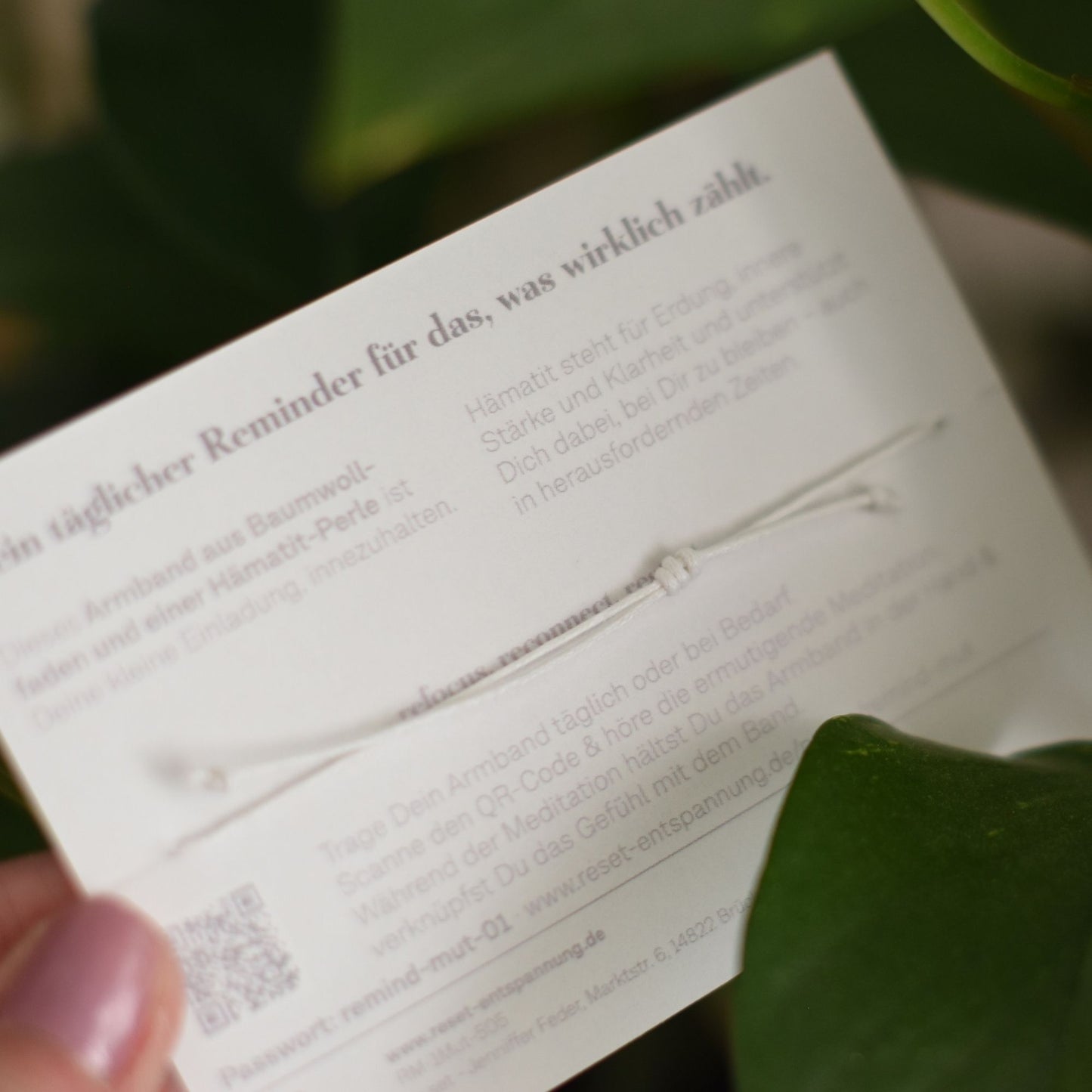 Nahaufnahme eines weißen Armbands mit Knoten auf einer Karte mit Text und QR-Code, gehalten von einer Hand vor grünem Blatt.