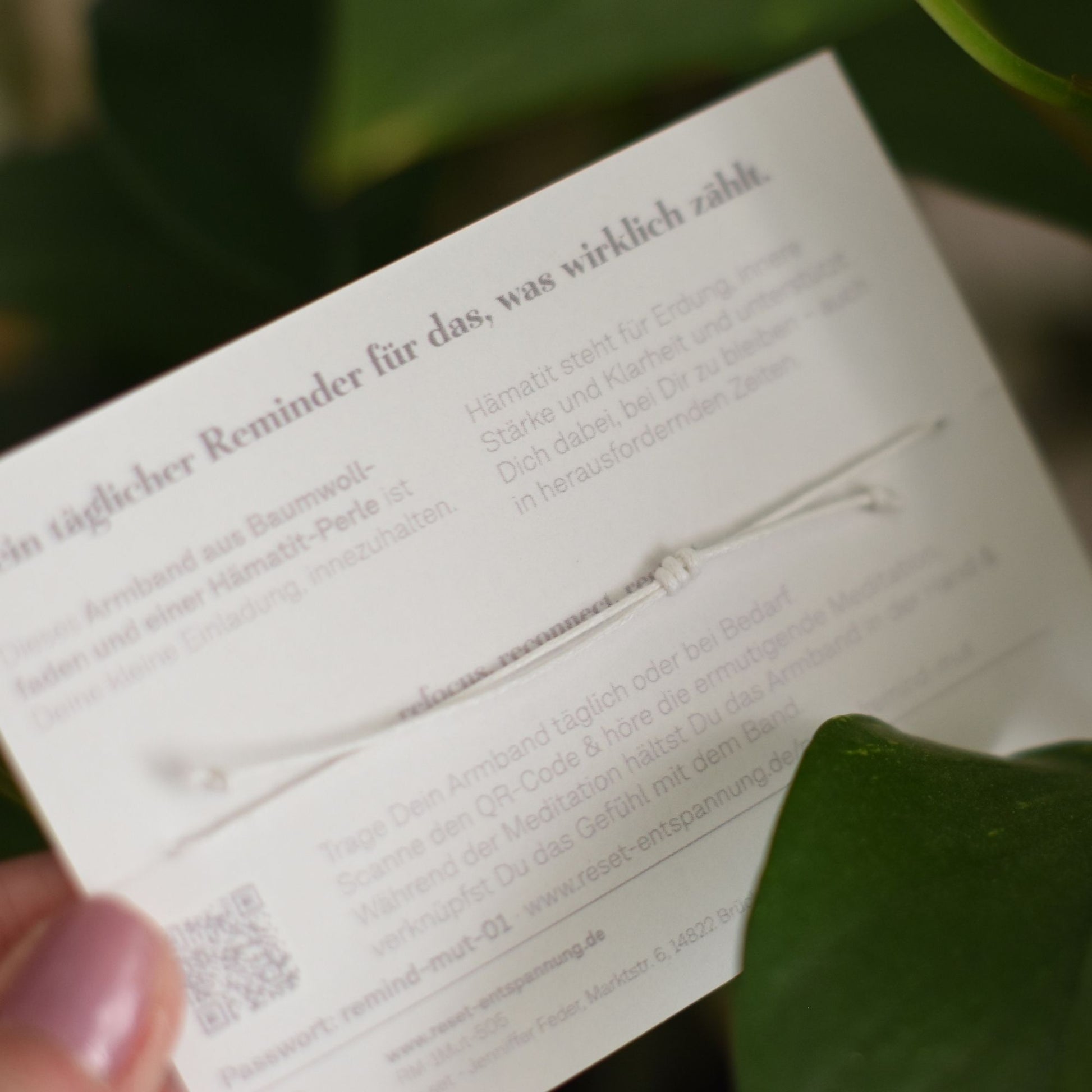 Nahaufnahme eines weißen Armbands mit Knoten auf einer Karte mit Text und QR-Code, gehalten von einer Hand vor grünem Blatt.