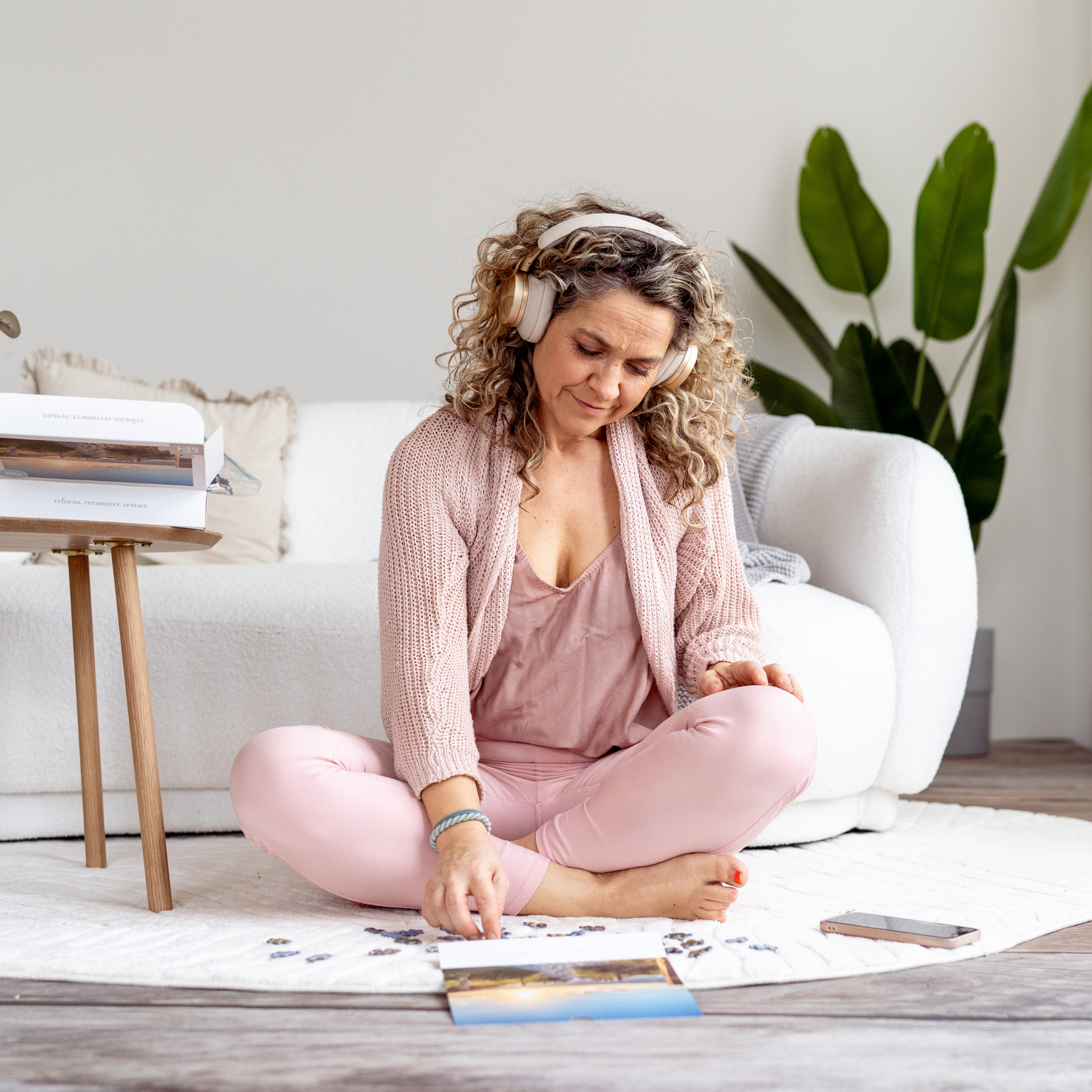 Frau sitzt im Schneidersitz auf einem Teppich vor einem Sofa und legt Puzzleteile auf eine Puzzlebox.