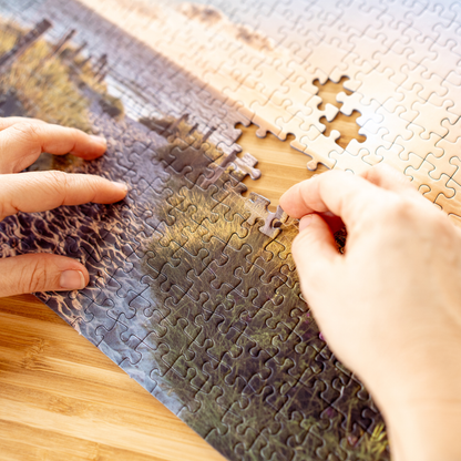 Hände fügen Puzzleteil in Landschaftsmotiv mit Wasser und Ufer ein, Puzzle auf Holzuntergrund.