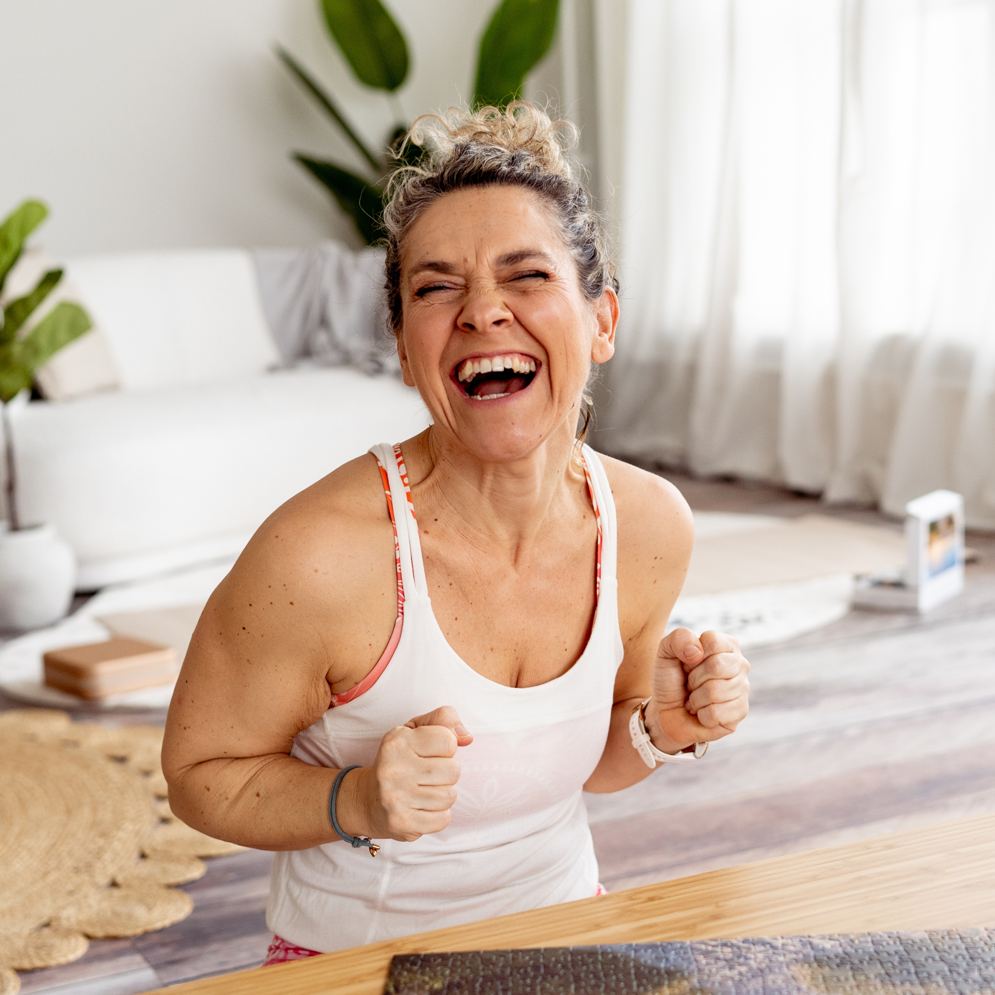 Frau in Sportkleidung sitzt an einem Tisch und freut sich sehr, im Hintergrund Pflanzen und helle Vorhänge.